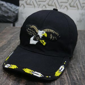 May include: Black baseball cap featuring a beaded eagle motif in white, yellow, and gold. The cap's brim is adorned with a beaded pattern in yellow, white, and black, offering a striking contrast.