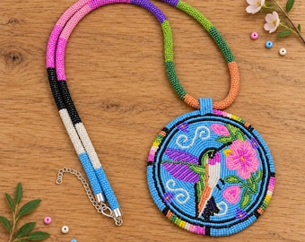 May include: A beaded necklace featuring a circular pendant with a hummingbird and flower design in pink, green, and blue. The cord is crafted from multi-colored beads in shades of pink, purple, green, orange, black, white, and blue with a metal chain.
