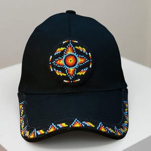 Może przedstawiać: Czarna czapka bejsbolowa z okrągłym, koralikowym wzorem pośrodku w kolorach pomarańczowym, żółtym, niebieskim i białym. Daszek ma również koralikową obwódkę w podobnych kolorach. Czapka znajduje się na białej powierzchni.