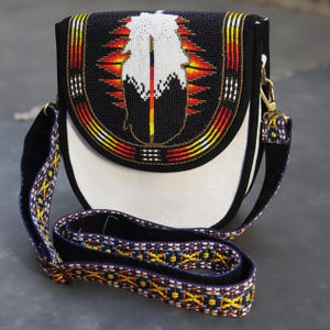 Pode incluir: Bolsa transversal preta e branca com um design de pena de contas. A bolsa apresenta uma base branca, uma aba preta com uma pena de contas colorida e uma alça estampada. A alça tem 101.6 cm de comprimento.