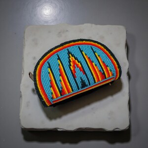 May include: A beaded, half-moon shaped pouch with a black base and zipper. The pouch features a turquoise background with a geometric design in red, orange, and yellow beads. The edges are outlined in black and red beads.