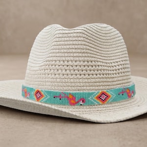 Puede incluir: Un sombrero fedora tejido de color crema con ala ancha y copa pellizcada. Una banda decorativa rodea la base, mostrando un fondo turquesa con un patrón repetitivo de flamencos rosas y formas geométricas de diamantes de colores.