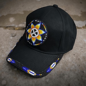 May include: Black baseball cap with a beaded design. The cap features a circular patch with a yellow and blue floral pattern and a white cross in the center. The brim is also decorated with a beaded pattern in blue, white, and yellow.
