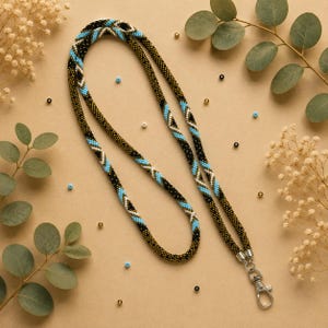 Może przedstawiać: Smycz z koralików z geometrycznym wzorem wykonanym z czarnych, turkusowych i białych koralików typu seed bead. Długi, rurowy sznurek jest zakończony u dołu srebrnym, metalowym karabińczykiem obrotowym, służącym do mocowania identyfikatorów lub kluczy.