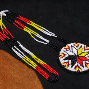 Puede incluir: Un collar de cuentas con un colgante circular con un diseño de estrella en negro, blanco, rojo y amarillo. Los hilos del collar son negros, blancos, amarillos y rojos. El collar se muestra sobre un fondo oscuro.