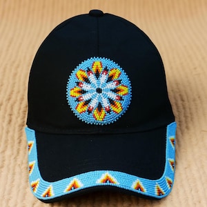 Può includere: Cappellino da baseball nero con un design di perline. Il cappellino presenta un emblema circolare di perline con un centro bianco, circondato da perline gialle, rosse e blu. La visiera è decorata con un motivo di perline blu.