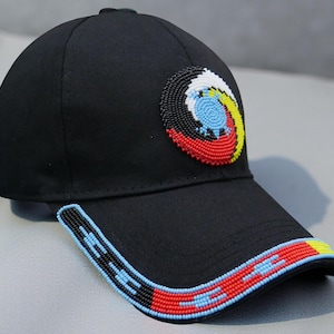 Può includere: Cappellino da baseball nero con un design di perline sulla parte anteriore e sulla visiera. Il design sulla parte anteriore è un motivo circolare con perline rosse, bianche, gialle e blu. La visiera ha un bordo di perline con perline rosse, blu e gialle.