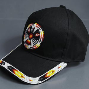 Może przedstawiać: Czarna czapka z daszkiem z wzorem z koralików. Przód zdobi okrągły wzór z czerwonych, żółtych i białych koralików. Daszek ma białą obwódkę z koralików z żółtymi i czerwonymi akcentami.