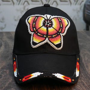 May include: Black baseball cap with a beaded butterfly design in shades of red, yellow, and orange. The brim of the cap is also decorated with a beaded pattern in red, white, and black. The cap is a stylish accessory.