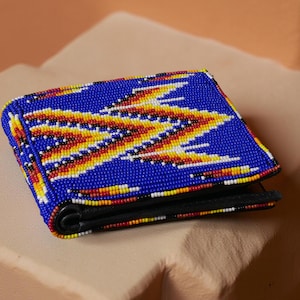May include: A blue beaded wallet with a geometric pattern in yellow, red, and white. The wallet is closed and has a black interior. The beaded design covers the entire front of the wallet.