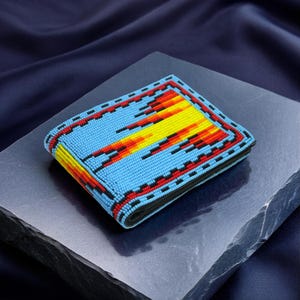May include: A beaded wallet with a vibrant design. The wallet features a light blue background with a geometric pattern in red, yellow, and orange. The edges are outlined in black and red. The wallet is displayed on a dark gray surface.