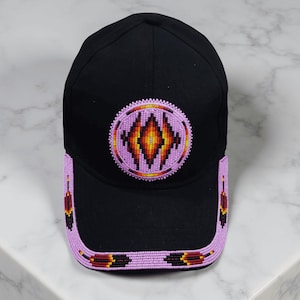 Könnte beinhalten: Schwarze Baseballkappe mit Perlendesign. Die Kappe hat ein kreisförmiges Design in der Mitte mit einem Rautenmuster in Orange-, Gelb- und Lilatönen. Der Rand und die Seiten sind mit einer Perlenborte in Lila, Rot und Gelb verziert.