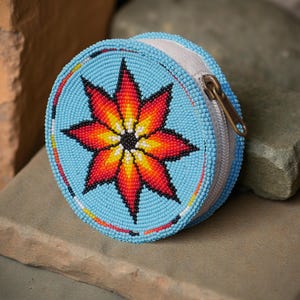 May include: Round, beaded coin purse in light blue with a vibrant, eight-pointed star design in red, orange, yellow, and black. The purse has a white zipper and a brass-colored zipper pull. The beaded design is a traditional Native American craft.