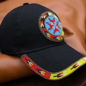 Puede incluir: Gorra de béisbol negra con un diseño de cuentas. La gorra presenta un emblema circular con cuentas rojas, azules y amarillas. La visera también está decorada con un patrón de cuentas en rojo, amarillo y blanco. La gorra está sobre un fondo marrón.