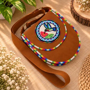 May include: A brown saddle-shaped crossbody bag featuring a circular beaded patch of a hummingbird feeding on a flower. The bag is accented with multi-colored beaded trim along the flap and edges, and includes an adjustable brown strap.
