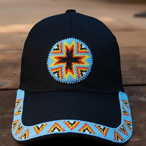 May include: Black baseball cap with a beaded design. The cap features a circular beaded emblem with a starburst pattern in orange, yellow, and black. The brim is also decorated with a beaded pattern in blue, orange, and white.