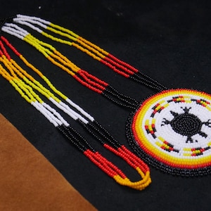 May include: A beaded necklace with a circular pendant featuring a black turtle design on a white background, surrounded by red, yellow, and black beads. The necklace strands are red, yellow, white, and black.