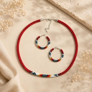 Pode incluir: Um conjunto de colar e brincos de argola feitos com missangas num padrão vibrante de vermelho, branco, preto, amarelo e turquesa. O colar inclui uma corrente extensora de metal prateado e um fecho de lagosta.
