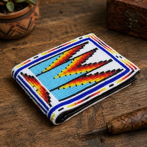 Peut inclure: Un portefeuille pliable orné de perles complexes sur l'extérieur. Le motif présente un fond blanc avec un dessin central de formes géométriques bleu clair, jaune, orange et rouge, encadré par une bordure de perles bleues, blanches et orange.