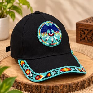 Puede incluir: Una gorra de béisbol negra con un parche circular de cuentas en la parte frontal que muestra un diseño de ave azul. La visera está decorada con un borde de cuentas continuo en patrones geométricos turquesa, rojos, amarillos y blancos.