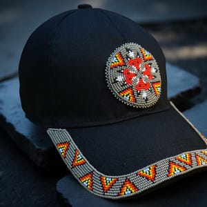 May include: Black baseball cap with a beaded design. The cap has a circular beaded patch with red, white, and grey beads. The brim is also decorated with a beaded pattern in orange, yellow, and blue. The cap is resting on a dark surface.