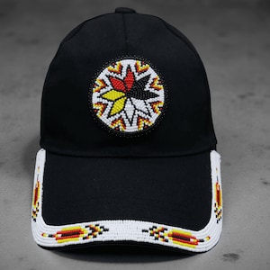 Op de afbeelding: Zwarte baseballcap met een kralenontwerp. De cap heeft een cirkelvormig embleem met een stervormig patroon in rood, geel en wit. De rand is ook versierd met een kralenpatroon in wit, rood, geel en zwart.