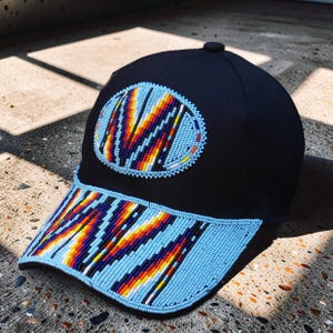 Op de afbeelding: Zwarte baseballpet met een kralenontwerp. De pet heeft een lichtblauwe, met kralen bezette klep en een cirkelvormige, met kralen bezette patch met een kleurrijk patroon van oranje, geel en blauw. De pet heeft een knoop aan de bovenkant.