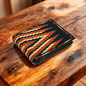 Pode incluir: Uma carteira preta com um padrão geométrico vibrante em vermelho, amarelo e branco. A carteira está dobrada e parece ser feita de um material tecido. O design assemelha-se a um padrão de chama ou seta estilizada.