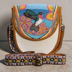 Könnte beinhalten: Umhängetasche mit weißem Korpus und einer Klappe mit einem aufgestickten Kolibri-Design. Die Klappe hat einen hellblauen Hintergrund mit bunten Blumen. Die Tasche hat eine braune Ledereinfassung und einen gemusterten Riemen.