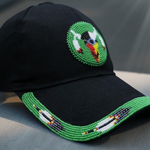 May include: Black baseball cap with a beaded design. The cap features a circular emblem with a green border and colorful accents. The brim is also adorned with a green beaded trim, adding a decorative touch to the hat.