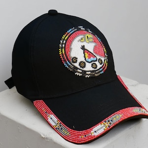 Könnte beinhalten: Schwarze Baseballkappe mit Perlendesign. Die Kappe hat ein kreisförmiges Emblem mit einem Vogel und geometrischen Formen in Rot, Weiß, Gelb und Blau. Der Rand ist mit roten, weißen und gelben Perlen verziert.