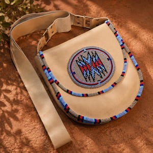 May include: A tan leather shoulder bag featuring a circular beaded medallion on the front flap. The medallion displays a geometric pattern in red, blue, black, and white. Two strands of multi-colored beads border the edge of the bag's flap.