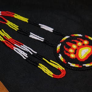 Pode incluir: Um colar de contas com um pingente redondo com um desenho de pata de urso. O colar e o pingente usam contas pretas, vermelhas, amarelas e brancas. O colar é exibido em uma superfície escura.