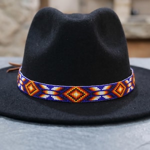 May include: A black fedora hat featuring a wide, flat brim and a pinched crown. The hat is accented with a decorative band made of small, colorful beads arranged in a geometric diamond pattern in shades of blue, orange, yellow, and white.