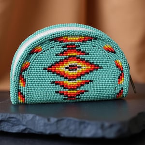 Può includere: Un portamonete di perline turchese con cerniera bianca e un motivo geometrico rosso, giallo e arancione. Il portamonete è a forma di mezzaluna e poggia su una superficie grigio scuro.