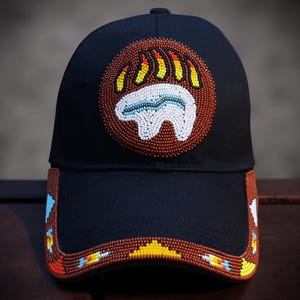 Op de afbeelding: Zwarte baseballcap met een kralen berenpootontwerp. De berenpoot heeft een cirkelvormig patroon met bruine, gele, witte en blauwe kralen. De rand van de pet is ook versierd met kleurrijke kralen.