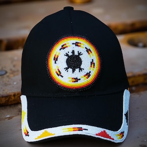 Pode incluir: Boné de beisebol preto com um design de contas. O boné apresenta um emblema circular com uma forma preta central rodeada por contas brancas, amarelas e vermelhas. A aba também é decorada com padrões de contas em cores semelhantes.