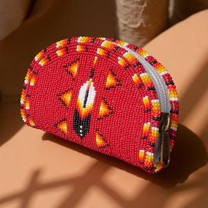 Könnte beinhalten: Eine rote, perlenbesetzte Geldbörse mit weißem Reißverschluss und einem bronzefarbenen Reißverschlussanhänger. Die Geldbörse hat ein zentrales Federdesign mit weißen, schwarzen, gelben und orangefarbenen Perlen und dreieckigen Mustern.