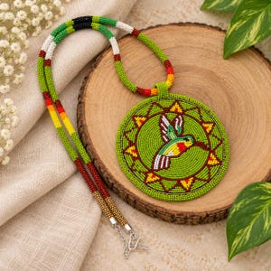 Peut inclure: Un collier pendentif circulaire en perles représentant un colibri vert au centre, entouré d'un motif géométrique en rayons de soleil rouge, jaune et blanc. Le long cordon en perles intègre des segments verts, noirs, blancs, rouges et jaunes.