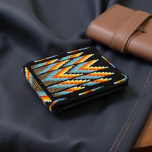 Puede incluir: Una cartera negra con cuentas con un diseño geométrico colorido en tonos de azul, naranja y amarillo. La cartera está doblada y parece estar hecha de cuero. Un estuche de cuero marrón está en el fondo.