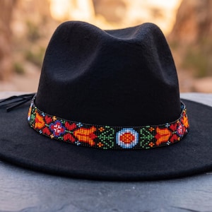 May include: A black fedora hat featuring a wide brim and a pinched crown. The hat is adorned with a decorative band made of small, colorful seed beads arranged in a repeating geometric pattern of red, orange, green, and blue floral motifs.