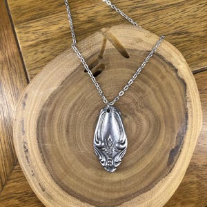 May include: A silver spoon pendant necklace with a decorative floral design. The spoon pendant hangs from a delicate silver chain. The necklace is displayed on a wooden surface.
