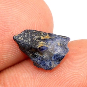 May include: A small, rough-cut sapphire gemstone. The sapphire is a deep blue color with areas of black and yellow. The gemstone is held between two fingers, with the skin visible in the background.