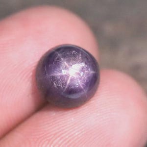 May include: A round, cabochon-cut star sapphire gemstone with a deep purple hue. The stone features a distinct six-rayed star effect, known as asterism, which appears as a bright, centred white light reflection on the polished, smooth surface.