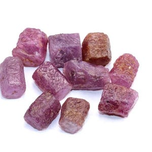 Puede incluir: Un conjunto de piedras preciosas de rubí en bruto, en varios tonos de rosa y rojo. Las piedras naturales tienen una superficie texturizada y están dispersas sobre un fondo blanco. Estas piedras son ideales para la fabricación de joyas o coleccionar.