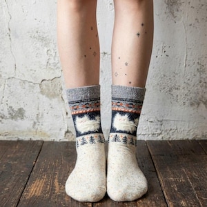 May include: Cream-coloured socks with a grey cuff and a patterned design. The socks feature a white cat face and a row of trees. The background is a wooden floor and a textured wall.