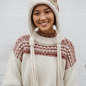 May include: A cream-colored knit hat with ear flaps and a decorative patch, paired with a matching sweater featuring red and cream horizontal stripes. The sweater has long tassels hanging from the neck.