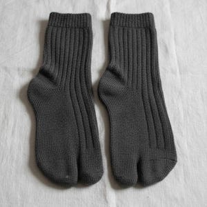 Puede incluir: Un par de calcetines tabi de color gris oscuro, dise&ntilde;ados con una puntera dividida. Los calcetines son acanalados y est&aacute;n hechos de un material de punto suave. Se muestran sobre una superficie blanca texturizada.