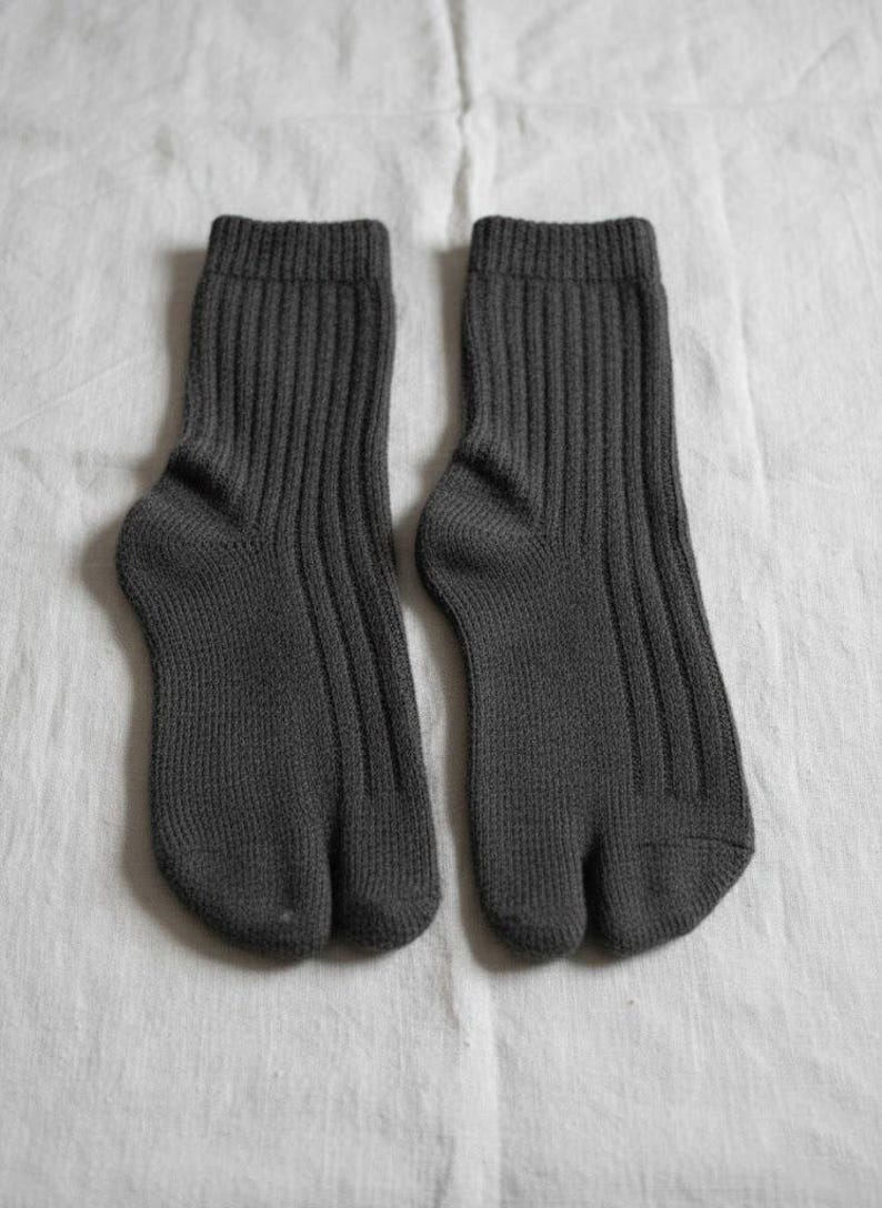 Puede incluir: Un par de calcetines tabi de color gris oscuro, dise&ntilde;ados con una puntera dividida. Los calcetines son acanalados y est&aacute;n hechos de un material de punto suave. Se muestran sobre una superficie blanca texturizada.