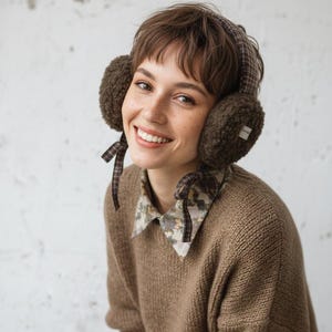 Puede incluir: Orejeras marrones con una diadema y orejeras a cuadros. Las orejeras tienen una textura suave y esponjosa y están atadas con cintas a cuadros. La persona lleva un suéter marrón y una camisa con cuello con un estampado floral.